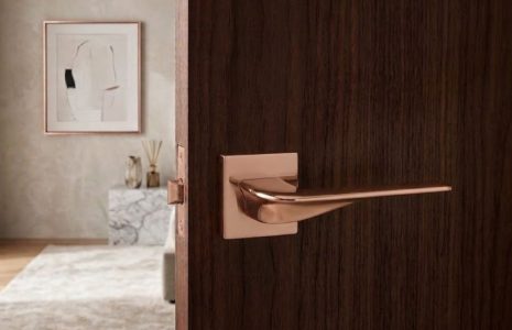 Close-up of a rose-gold door handle on a dark wood door, opening to a bright, minimalist living room with marble table and abstract art.